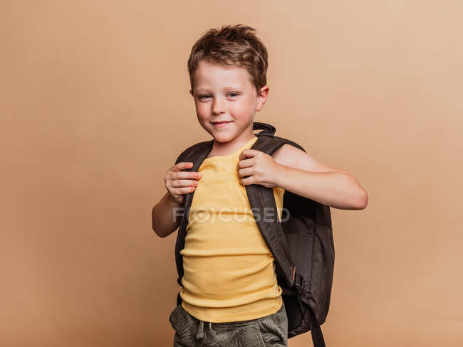 Позитивний крутий дев'ятнадцятикласник з рюкзаком дивиться на камеру на коричневому фоні в студії — стокове фото