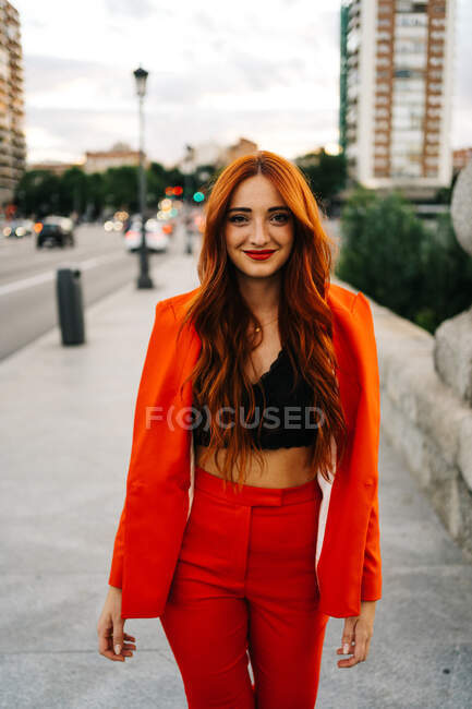 Affascinante femmina con lunghi capelli rossi e in abito arancione alla moda in piedi in strada la sera e guardando la fotocamera — Foto stock
