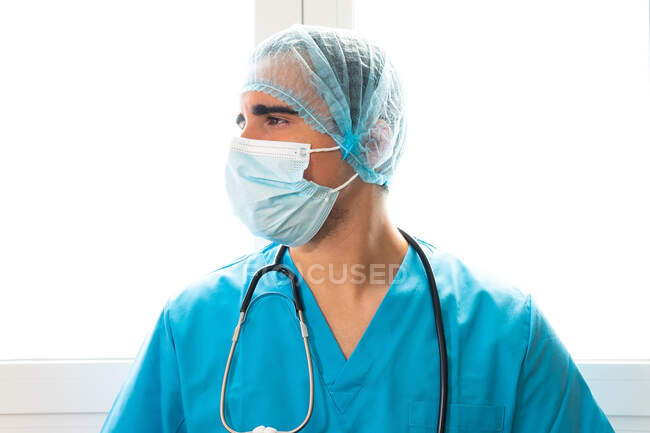 Vista lateral do médico masculino em máscara e uniforme de pé perto da janela na clínica — Fotografia de Stock