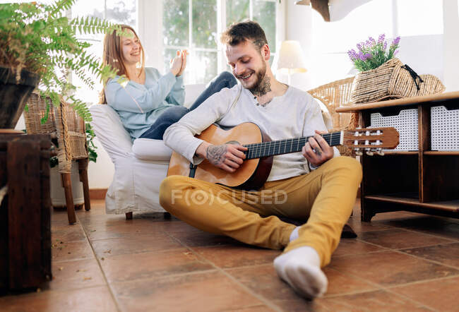 Musicien masculin tatoué aux jambes croisées jouant de la guitare acoustique contre des applaudissements féminins joyeuses dans un fauteuil à la maison — Photo de stock