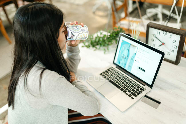 High angle side view of female buyer drinking beverage while sitting at table with laptop and choosing goods during online shopping — Stock Photo