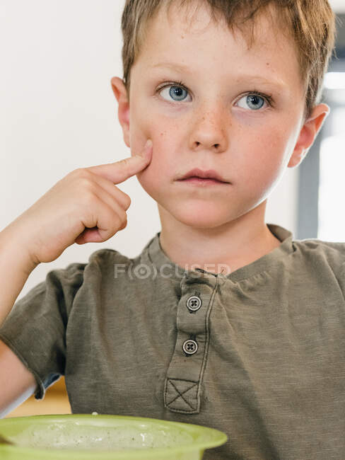 Adorable child touching cheek while sitting at table with bowl of food during lunch and looking away — Stock Photo