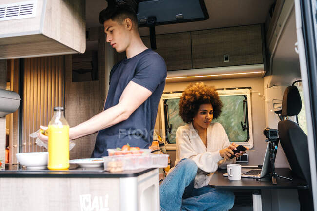 Jovem preparando café da manhã saudável, enquanto a namorada afro-americana usando gadgets durante a viagem de verão juntos em van campista — Fotografia de Stock
