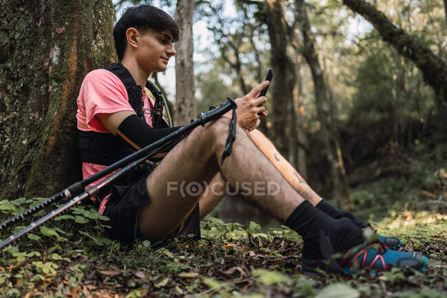 Side view of content male hiker with backpack sitting in woods and browsing mobile phone while traveling — Stock Photo