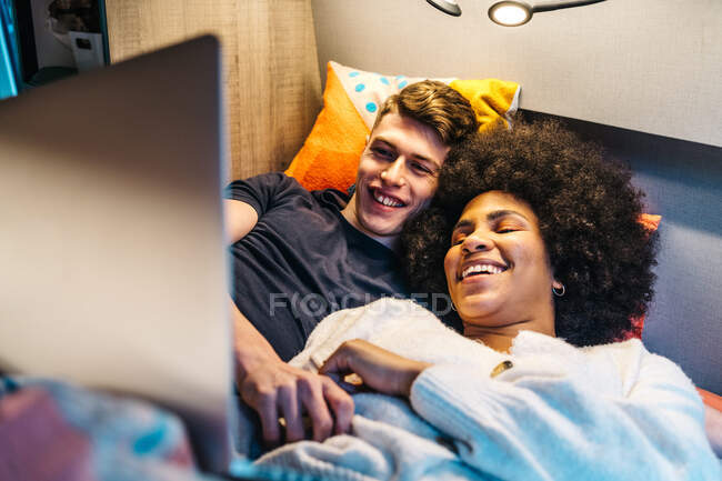 Glückliches romantisches multiethnisches Paar, das kuschelt und die Zeit zusammen genießt, während es im Bett liegt und während der Reise im Wohnmobil Filme auf dem Laptop anschaut — Stockfoto