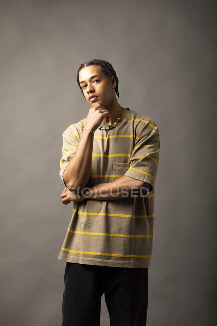 Молодой вдумчивый афроамериканец с плетеными волосами, одетый в широкую полосатую рубашку и ожерелье, смотрит в камеру на сером фоне — стоковое фото