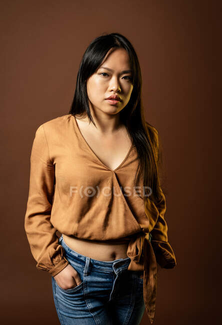 Vue de face de la femelle asiatique en vêtements à la mode regardant la caméra sur fond brun en studio — Photo de stock