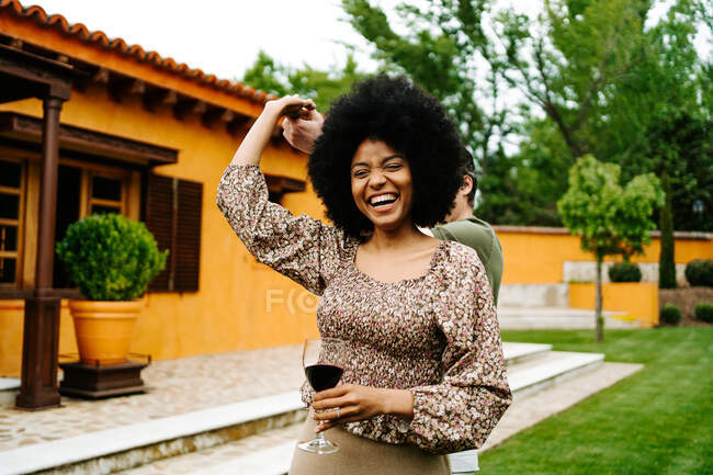 Contenu femme noire avec verre de vin rouge tenant la main de l'homme et dansant dans la cour près du chalet tout en s'amusant — Photo de stock
