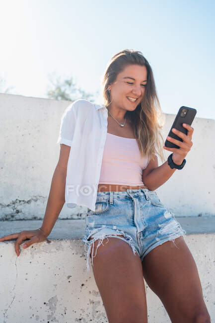 Positive cheerful young female browsing on mobile phone on sunny day in summer in city — Stock Photo
