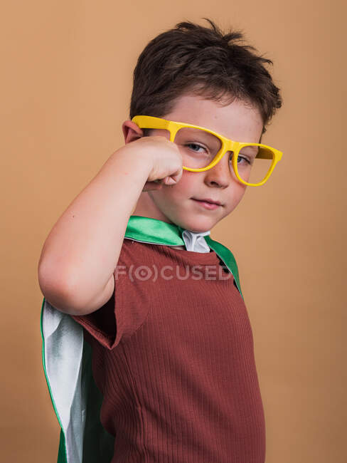 Вид збоку дитини в супергеройському мисі і декоративні окуляри, що показують жест міцності при погляді на камеру — стокове фото