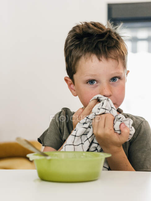 Милый мальчик вытирает рот салфеткой, сидя за столом с миской сливочного супа во время обеда дома — стоковое фото