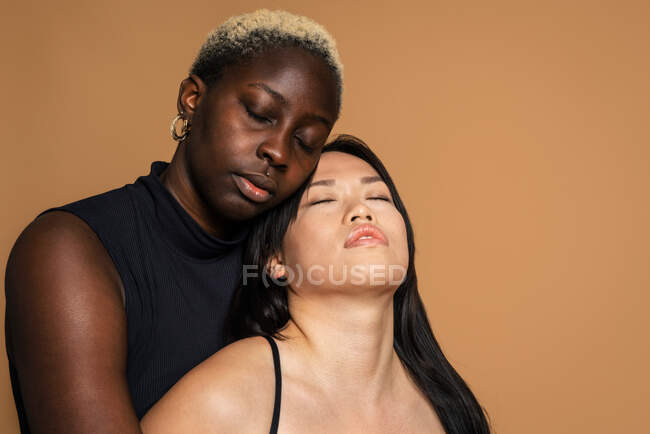 Modèles féminins multiraciaux en lingerie noire étreignant sur fond beige pour concept de positivité corporelle en studio les yeux fermés — Photo de stock