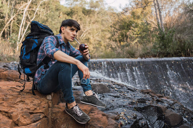 Durstige männliche Wanderer mit Rucksack trinken Wasser aus Becher mit Stroh, während sie auf einem Felsen in der Nähe des Wasserfalls im Wald sitzen und in der Pause wegschauen — Stockfoto