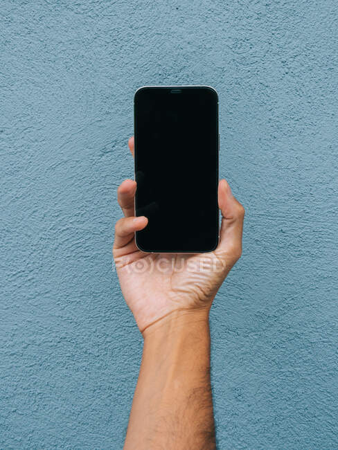 Recorte masculino irreconocible que muestra el teléfono móvil moderno con pantalla negra sobre fondo azul en la ciudad - foto de stock