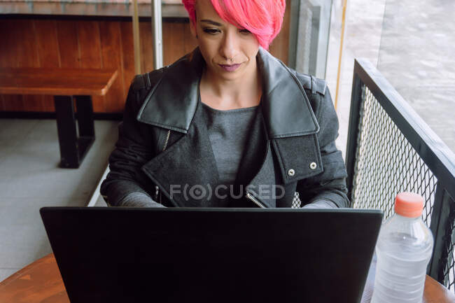 Зверху жінка з фарбованим волоссям у стильному пальто, що сидить за столом біля вікна при використанні ноутбука — стокове фото