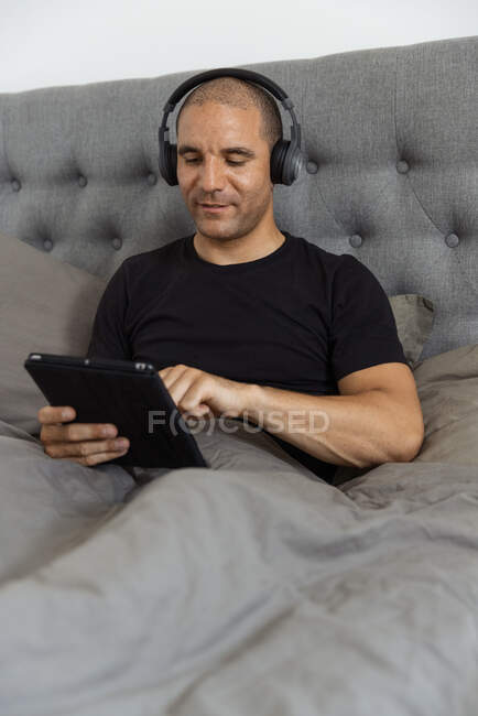 Gelassener Mann mit Kopfhörer sitzt auf dem Bett unter einer Decke und surft auf dem Tablet in den sozialen Medien, während er morgens Musik hört — Stockfoto