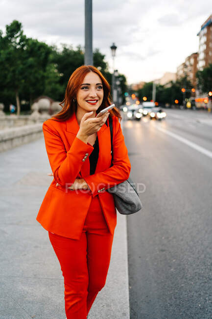 Smiling female with red hair and in orange suit recording audio message on mobile phone while communicating with friend on social media and standing in city street — Stock Photo