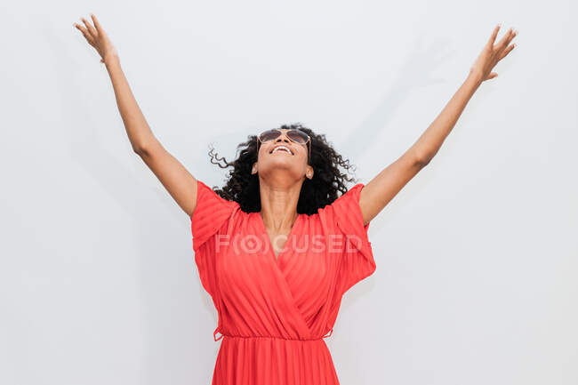 Захоплена афро - американка в червоному одязі і сонцезахисних окулярах, дивлячись вгору з піднятими руками на світлому тлі — стокове фото