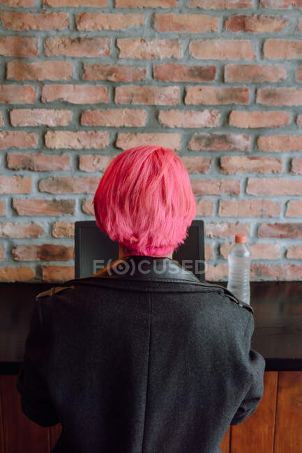 Voltar ver mulher colheita com cabelo tingido no casaco elegante sentado à mesa perto da parede de tijolo ao usar laptop — Fotografia de Stock