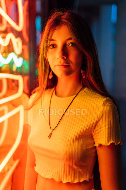 Junge sanfte Frau mit Anhänger und Ohrring mit langen Haaren blickt in die Kamera, während sie abends von orangefarbenen Neonlichtern beleuchtet wird — Stockfoto