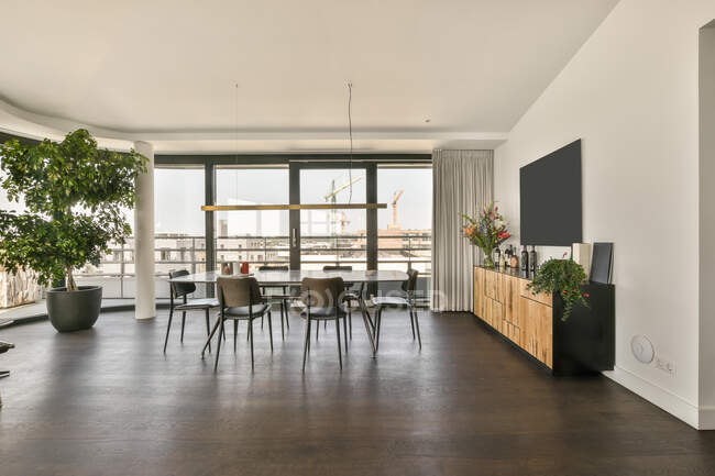Mesa de jantar e cadeiras colocadas perto de grande janela panorâmica em quarto espaçoso com TV e armário em apartamento estilo loft moderno — Fotografia de Stock