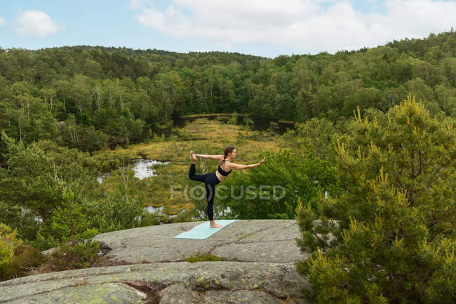Vue latérale femelle mince faisant Parivritta Natarajasana pose sur le rocher lors d'une séance de yoga le jour d'été dans la nature verte — Photo de stock
