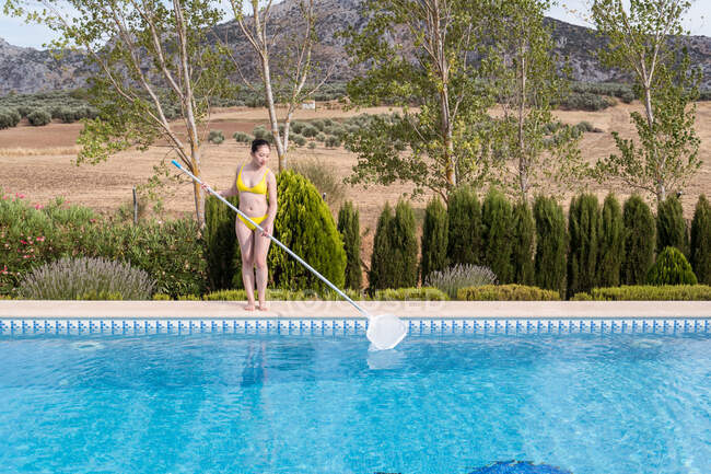Mulher étnica em biquíni limpeza de água da piscina com rede no dia ensolarado no verão — Fotografia de Stock