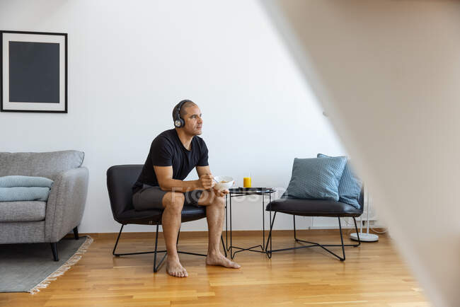 Hombre pacífico comiendo deliciosa comida para el desayuno mientras escucha música en los auriculares y sentado en la silla en el salón en casa - foto de stock