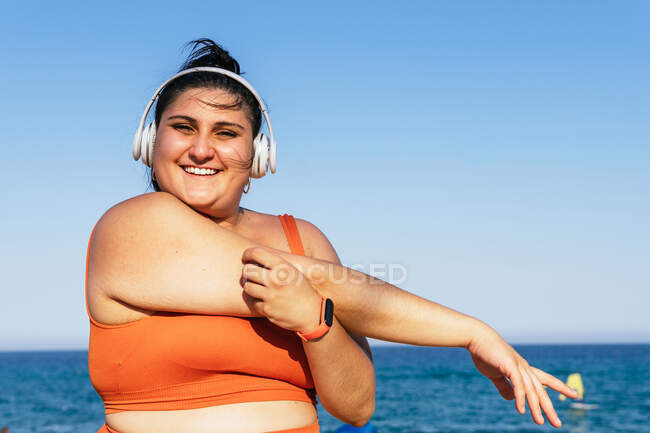 Atleta feminina étnica alegre com corpo curvilíneo ouvindo música de fones de ouvido enquanto olha para a câmera e se estende sob um céu azul — Fotografia de Stock