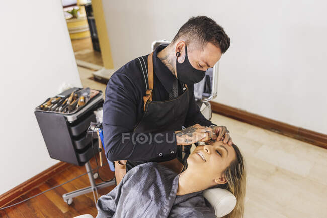 D'en haut l'homme dans le masque enduisant fondation du visage de la femme blonde pendant le travail dans le studio de maquillage professionnel — Photo de stock