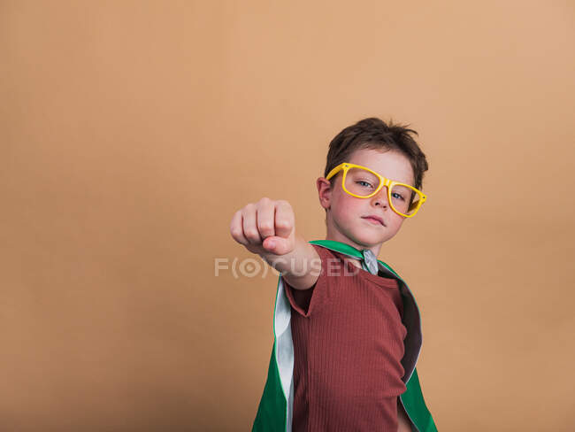 Bambino con mantello da supereroe e occhiali decorativi che mostrano un gesto di forza mentre guarda la fotocamera — Foto stock