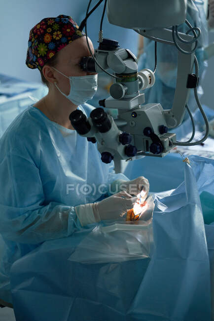 Médica atenta em uniforme cirúrgico e máscara estéril olhando através do microscópio enquanto operava o olho de paciente irreconhecível no hospital — Fotografia de Stock