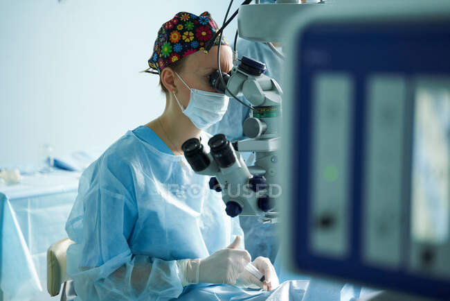 Уважний лікар-жінка в хірургічній формі та стерильній масці, яка дивиться через мікроскоп під час операції очей невпізнаваного пацієнта в лікарні — стокове фото