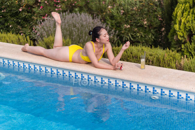 Vista lateral de la hembra asiática en bikini amarillo tumbada cerca de la piscina y tomando el sol mientras disfruta de las vacaciones de verano en el resort - foto de stock