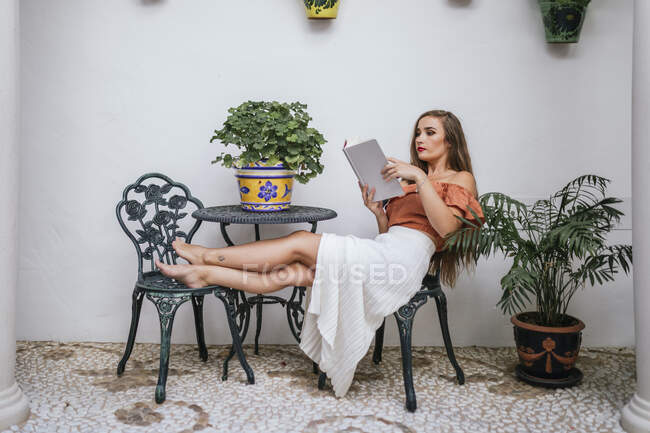 Vista laterale di graziosa femmina in gonna gambe appoggiate sulla sedia mentre si rilassa in patio in estate e godersi la storia interessante nel libro durante il fine settimana — Foto stock