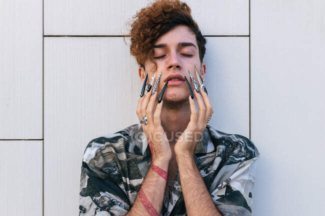 Молодой тщеславный мужчина в стильной одежде с длинными ногтями, стоящими на черепичной стене с закрытыми глазами — стоковое фото