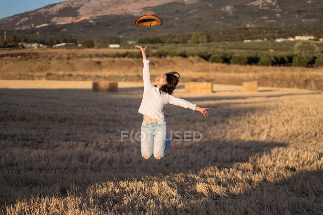 Жінка кидає капелюх і стрибає по сухому полі в літній вечір у сільській місцевості — стокове фото