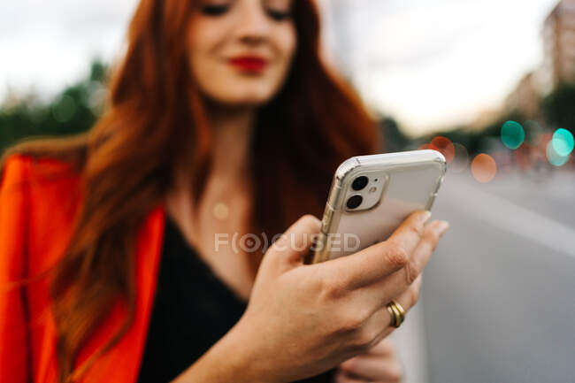 Mulher sorridente com cabelo vermelho e em mensagens de texto de terno laranja no telefone celular enquanto caminha na rua da cidade — Fotografia de Stock