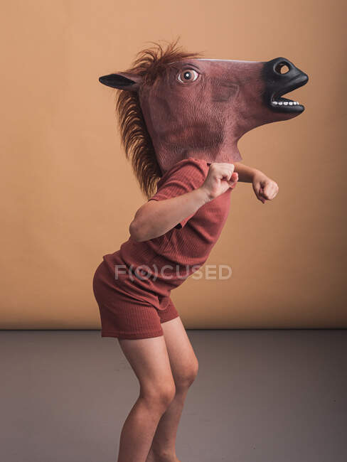 Seitenansicht eines anonymen Kindes in Pferdemaske, das sich nach vorne lehnt, während es galoppierende Hengste auf beigem Hintergrund darstellt — Stockfoto