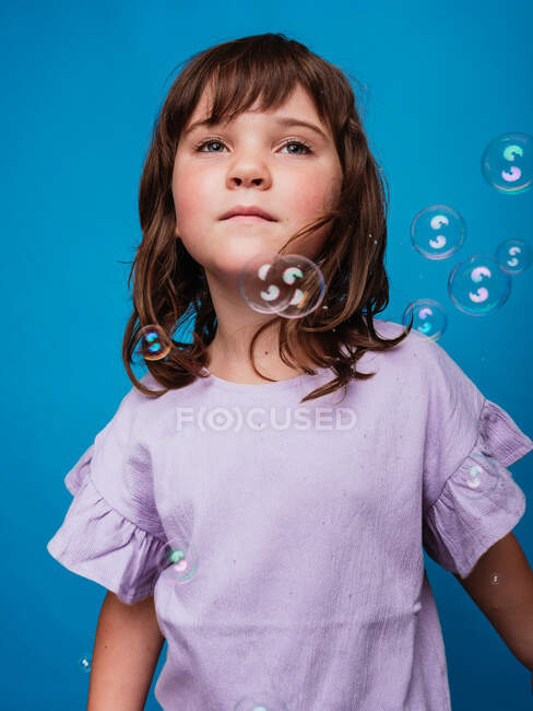 Безтурботний дитина в сукні стоїть в студії з плаваючою мильною бульбашкою на яскраво-блакитному фоні — стокове фото