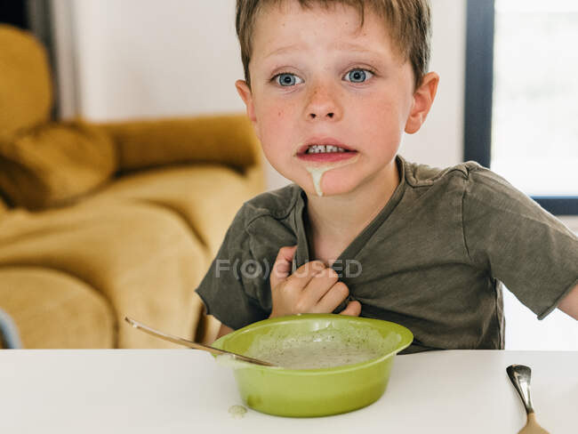 Menino com boca suja sentado à mesa com tigela de sopa de creme durante o almoço e olhando para longe — Fotografia de Stock