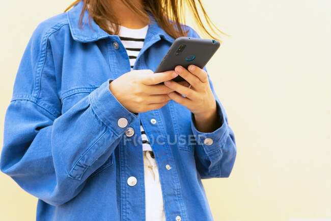 Ritagliato irriconoscibile giovane donna in abito alla moda messaggistica sul telefono cellulare sullo sfondo del muro in strada della città e guardando altrove — Foto stock