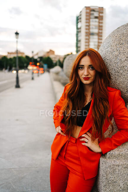 Приваблива жінка з довгим рудим волоссям і в модному помаранчевому костюмі стоїть на вулиці ввечері і дивиться на камеру — Stock Photo