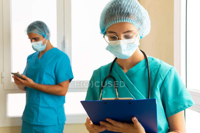 Médica mulher em relatório de leitura uniforme médica na área de transferência, enquanto em pé no hospital no fundo do médico masculino desfocado — Fotografia de Stock