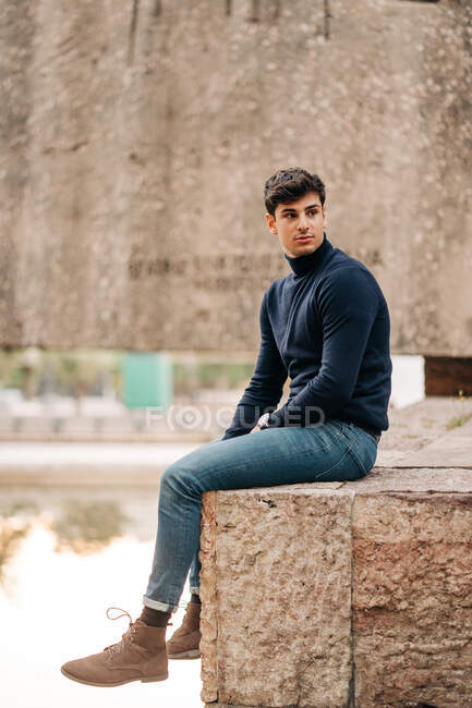 Side view of young male in trendy outfit sitting on stone fence in city and looking away — Stock Photo