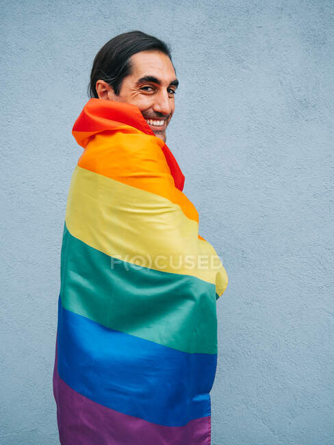 Gay ethnic male wrapped in rainbow LGBT flag looking at camera against gray wall in city — Stock Photo