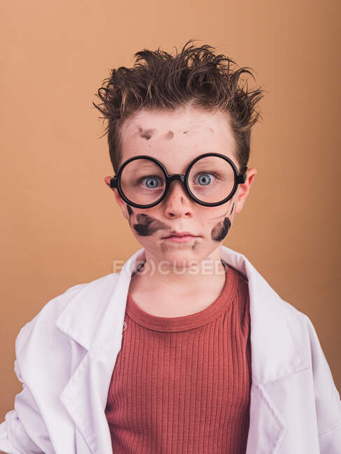 Químico menino em roupão de laboratório e óculos de plástico olhando para a câmera com olhar louco no fundo bege — Fotografia de Stock