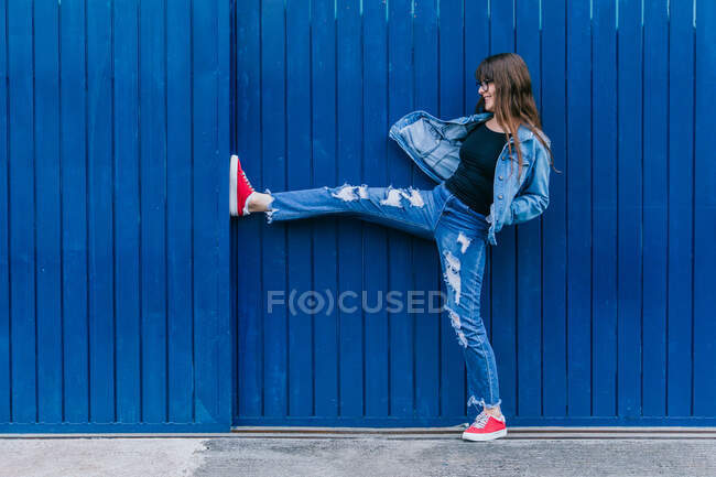 Sorrindo hipster feminino em roupas de ganga inclinando-se na parede azul na rua da cidade — Fotografia de Stock