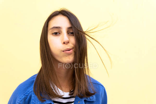 Молода жінка регулює коричневе волосся повітряним ударом на пастельному жовтому тлі і дивиться на камеру — стокове фото