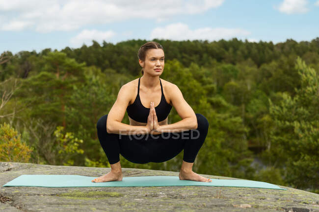 Mujer joven descalza de cuerpo entero haciendo pose de Malasana en la esterilla y mirando hacia otro lado mientras practica yoga sobre roca en la naturaleza en verano - foto de stock